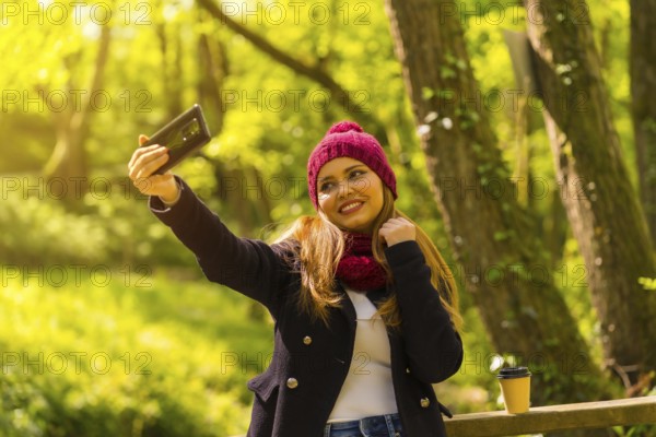 Young man with a black jacket, scarf and red woolen hat enjoying in an autumn park, taking a selfie