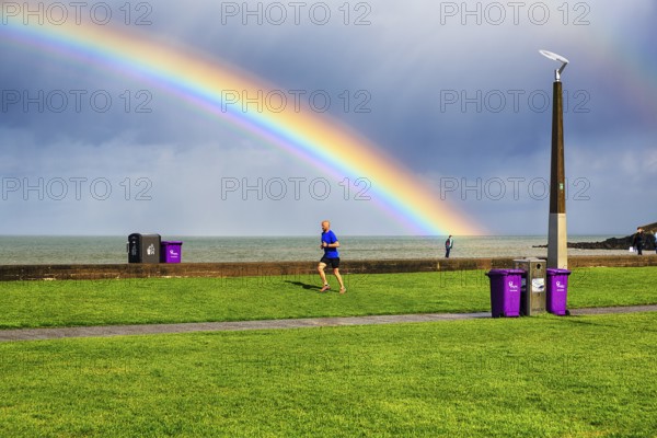 Pedestrians and joggers on seafront promenade, dramatic cloudy sky with rainbow, Bray, Wicklow,