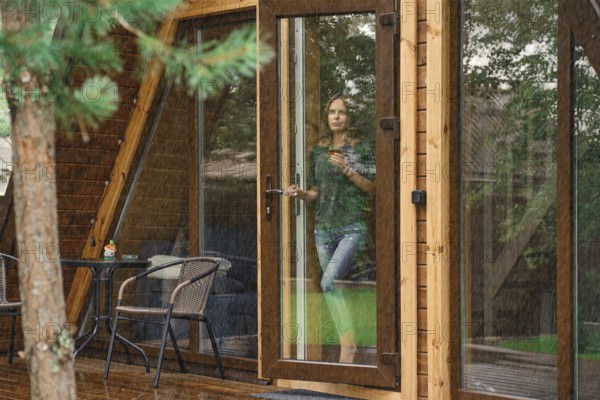 Young woman with a cup of herbal tea stands on the doorstep of a forest cabin and looks at the rain