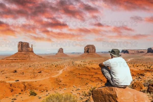 A young boy with white t-shirt sitting in the center of the photo on a stone in the Monument Valley