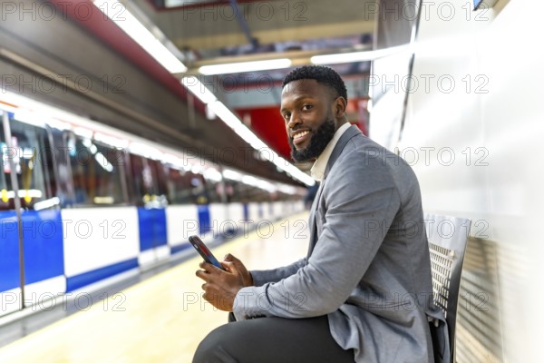 Young businessman is sitting on bench in subway station and using smartphone while waiting for
