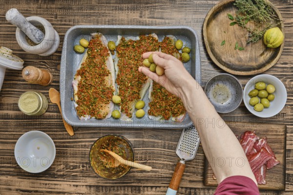 A hand adds green olives to a tray of seasoned fish fillets covered in vibrant herbs and spices.