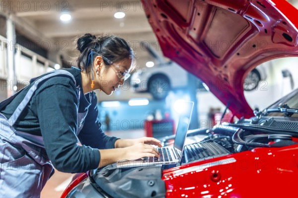 Young female mechanic using a laptop while performing diagnostics on a red car in a modern auto