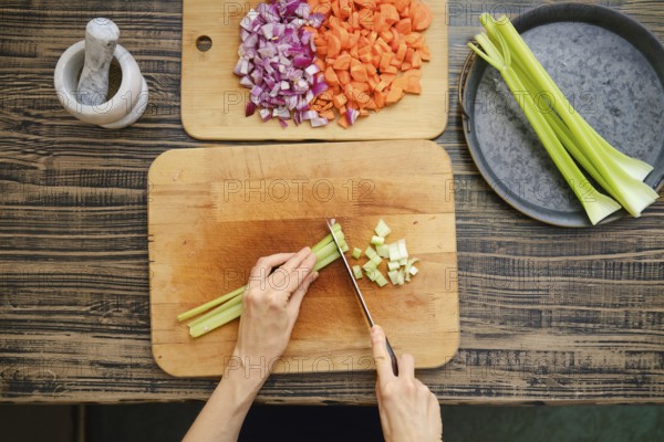 A person is chopping celery on a wooden cutting board, surrounded by diced onions and carrots. The