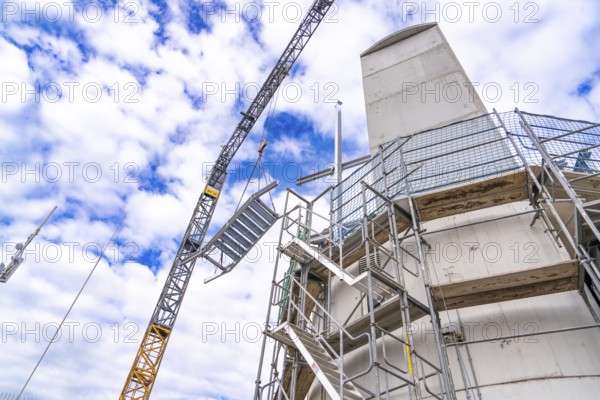 Construction crane lifts staircase structure next to tower and scaffolding in front of a cloudy