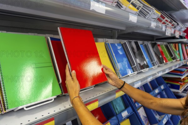 A person is holding a green spiral notebook in a store. There are many notebooks on the shelves,
