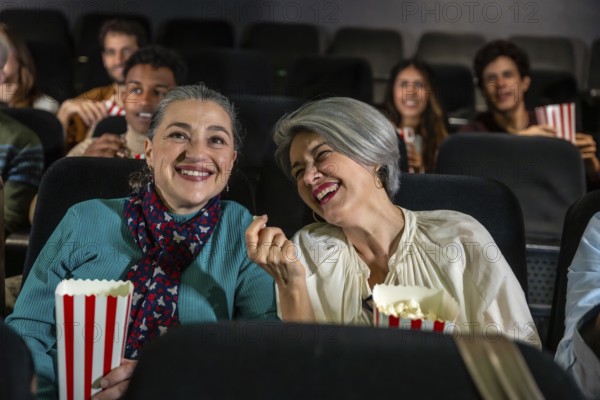 Two cheerful senior women laughing and eating popcorn while watching a movie at the cinema, sharing