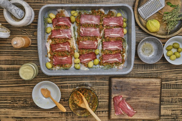 Fresh fish fillets layered with bacon and seasoning, herbs, and olives sits on a baking tray.