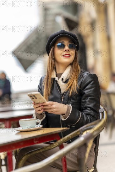Young woman wearing sunglasses and a hat using mobile phone while sitting at a table in a street