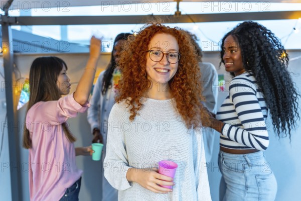 Young woman with redhead curly hair and eyeglasses smiling standing with beer glass in a roof party