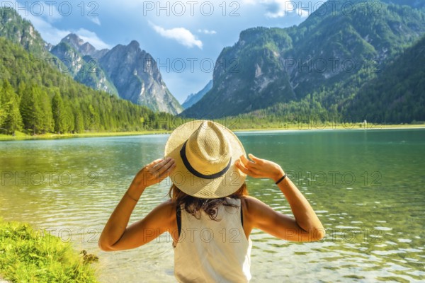 Young woman holding her straw hat admiring the landscape of lake dobbiaco and the dolomites during