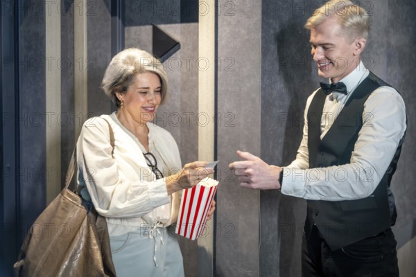 Elegant mature woman handing ticket to smiling usher while holding popcorn bucket, ready to enjoy