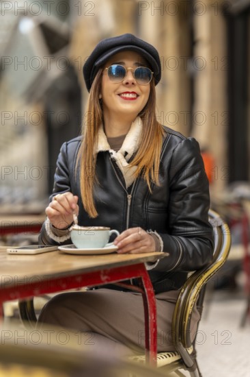 Young woman wearing sunglasses and baker boy hat stirring cappuccino at outdoor cafe table with