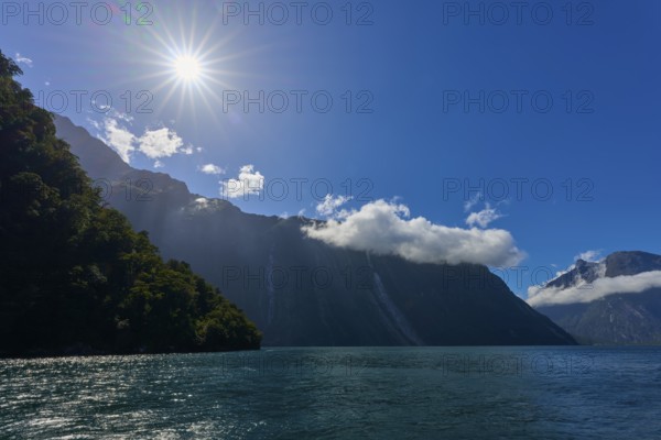 Sunbeams shine over majestic mountains and a calm lake under a clear sky, summer, Milford Sound, Te