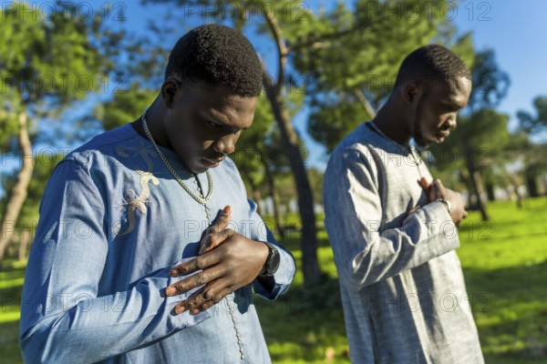 Two young senegalese men wearing traditional dashiki clothing are praying outdoors in a park,