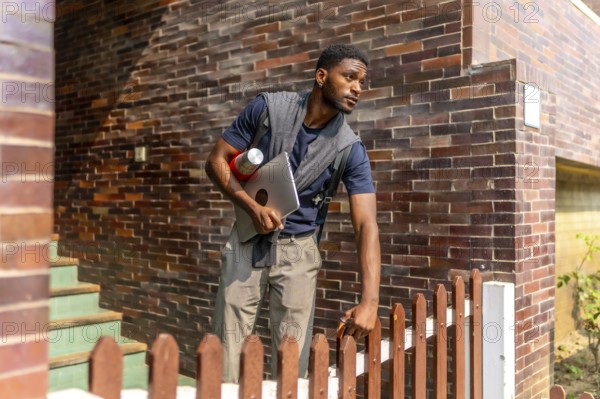 Young african businessman leaving a house in a residential district to go to work holding a laptop