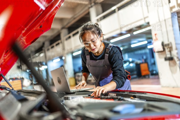 Young woman working as a mechanic, using a laptop while inspecting the engine of a red car in a