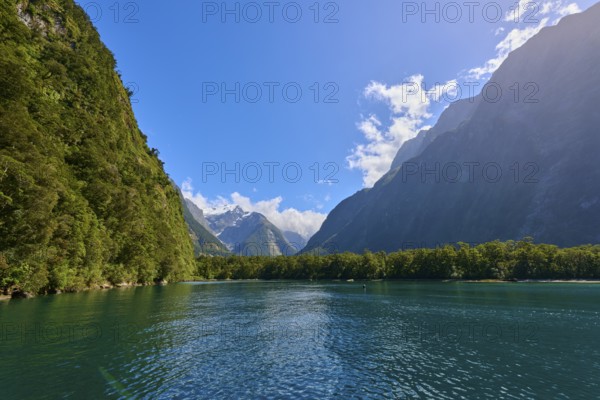 Calm waters between forested slopes and towering mountains under a clear sky, fjord landscape,