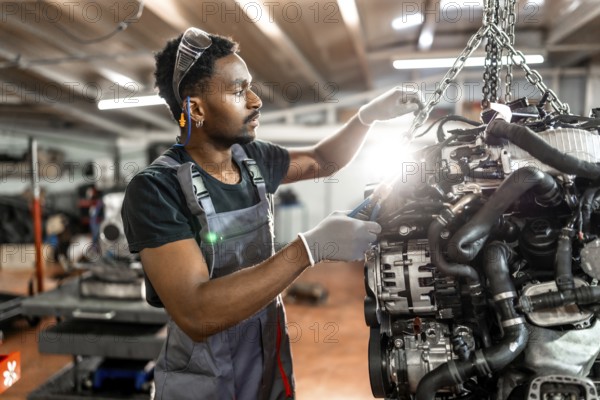 Young mechanic wearing protective gear working on a car engine suspended by a chain hoist in a