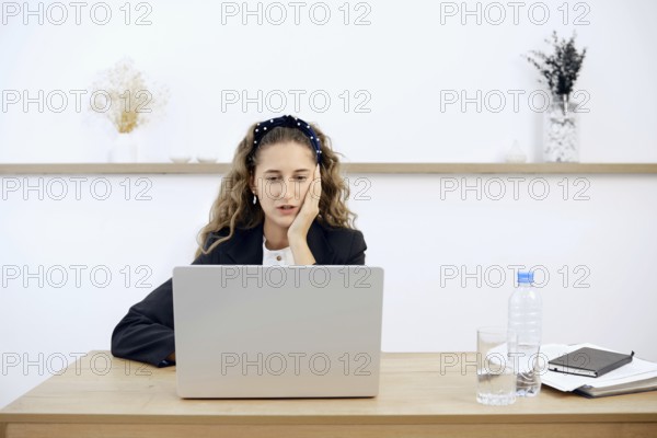 A young woman sits at a wooden desk, focused on her laptop while resting her chin on her hand. A