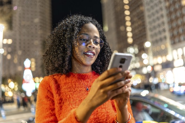 Young woman wearing an orange sweater, smiling while using a smartphone on a bustling city street,
