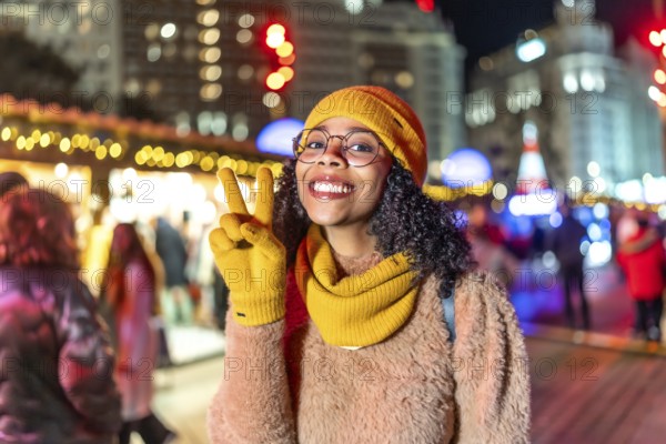 Smiling young woman wearing yellow gloves, hat and scarf showing peace sign while enjoying