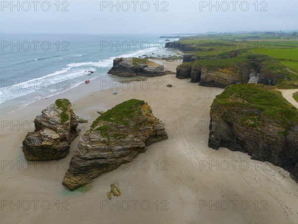 Wide beach with scattered large rock formations and calm water, bordered by steep coastal cliffs,