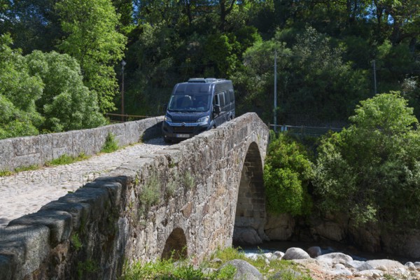 A van drives over a historic stone bridge, surrounded by lush vegetation and sunny weather,