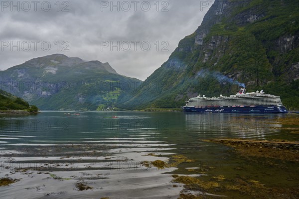 A large cruise ship, Mein Schiff 6, lies in the fjord, surrounded by green mountains under a cloudy