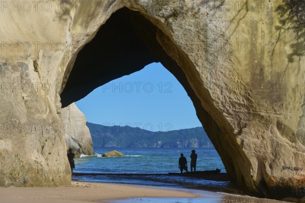 Silhouettes of people under a large rock arch on the beach overlooking the sea, Cathedral Cove,