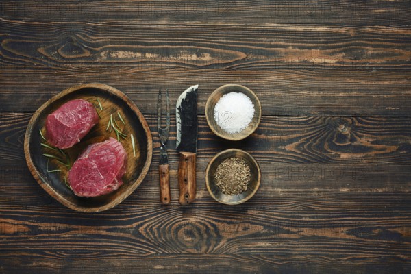 Two raw steaks on a wooden plate alongside bowls of salt and pepper, with old forged knife and fork