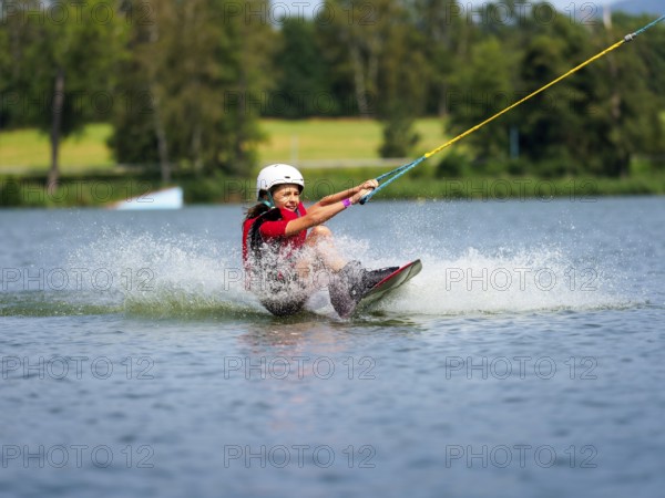 Boy or girl, sitting on wakeboard, cheerful, red life jacket, water skiing and summer holiday in