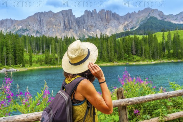 Woman with straw hat and backpack admiring the turquoise waters of lake carezza with the latemar