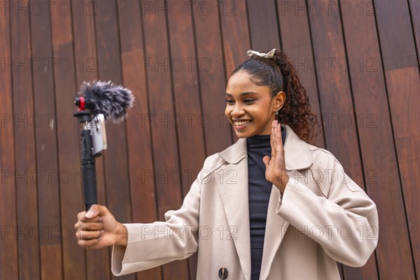 Young african american woman influencer waving at camera while recording vlog outdoors next to a