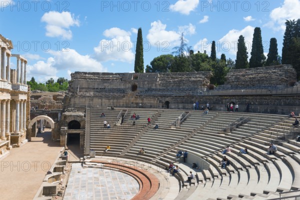 An ancient amphitheatre with stone seats under a blue sky with clouds and trees in the background,