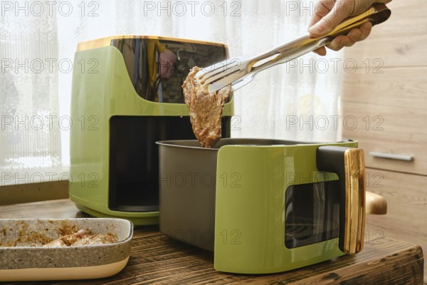 A hand holds tongs putting a piece of marinated meat to an air fryer. The process of preparing