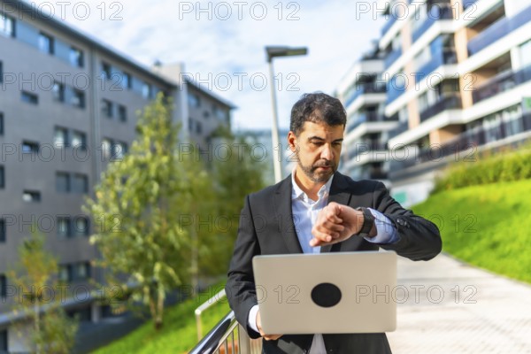 Mature adult caucasian businessman checking time to smartwatch while standing using laptop at city