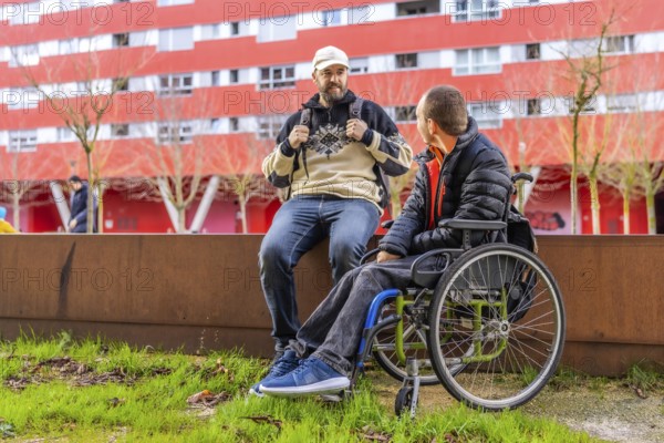 Two male friends, one using a wheelchair, are sitting on a low wall, chatting and enjoying their