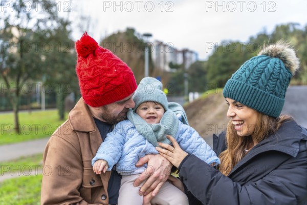 Parents holding their baby while smiling and enjoying a joyful winter day in a park, surrounded by