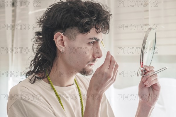 Concentrated young man with curly hair plucking his eyebrows with tweezers while looking in a small