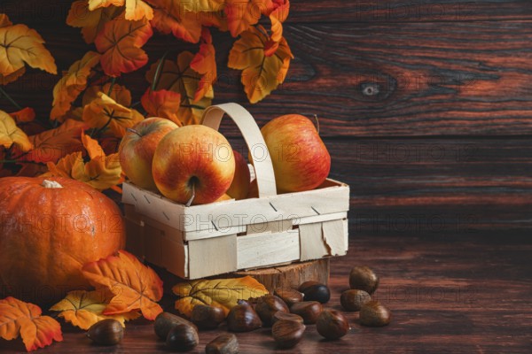 A wooden basket with apples next to chestnuts, a pumpkin and autumn leaves