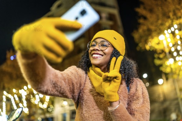 Smiling woman wearing yellow winter clothes taking selfie with smartphone and making peace sign in