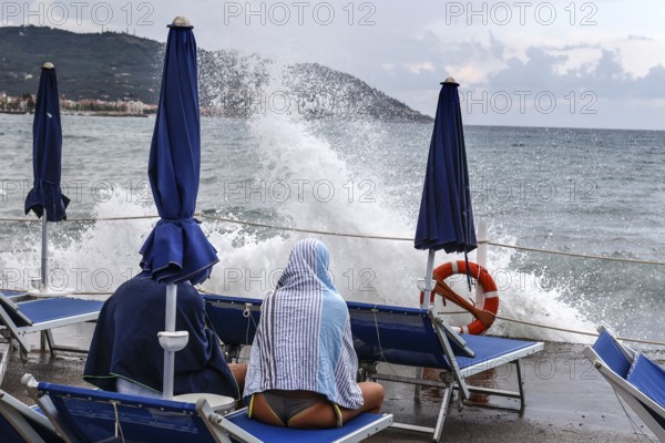Waves crashing against the pier of a bathing jetty where a couple is sitting on deckchairs, Diana