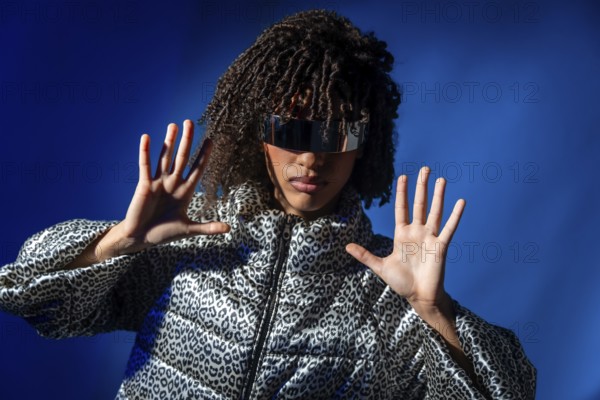 Stylish model wearing futuristic visor and patterned jacket pushing with hands on vibrant blue