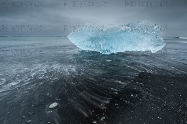 A shimmering blue iceberg is washed up by waves on a dark beach, Diamond Beach, Jökulsárlón glacier