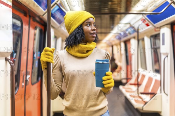 Young black woman wearing yellow winter clothes, holding a reusable coffee cup and traveling on a