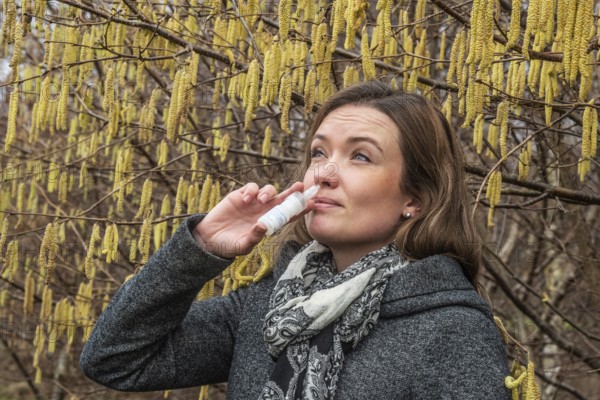 Young Caucasian woman with allergy problem stands by flowering hazel bush and sprays her nose in