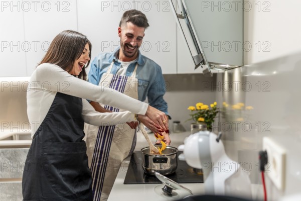 Happy couple wearing aprons preparing food in modern kitchen, stirring in pot and laughing
