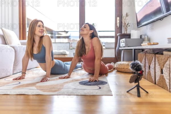 Two women recording an online yoga class from home, performing the cobra pose on a rug in the