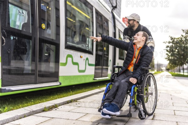 Young man in a wheelchair points at an approaching tram while his friend stands behind him,